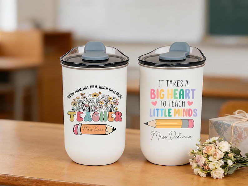 May include: Two white travel mugs with black lids and gray flip-top closures. One mug has the text "Teach them, love them, watch them grow TEACHER" with a pencil graphic. The other mug says "It takes a big heart to teach little minds."