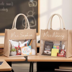 May include: Two beige tote bags with clear front pockets, filled with gifts. One bag says "Mrs. Sophia" and the other says "Mrs. Nancy" in white script. The bags sit on a wooden desk, with a chalkboard in the background.