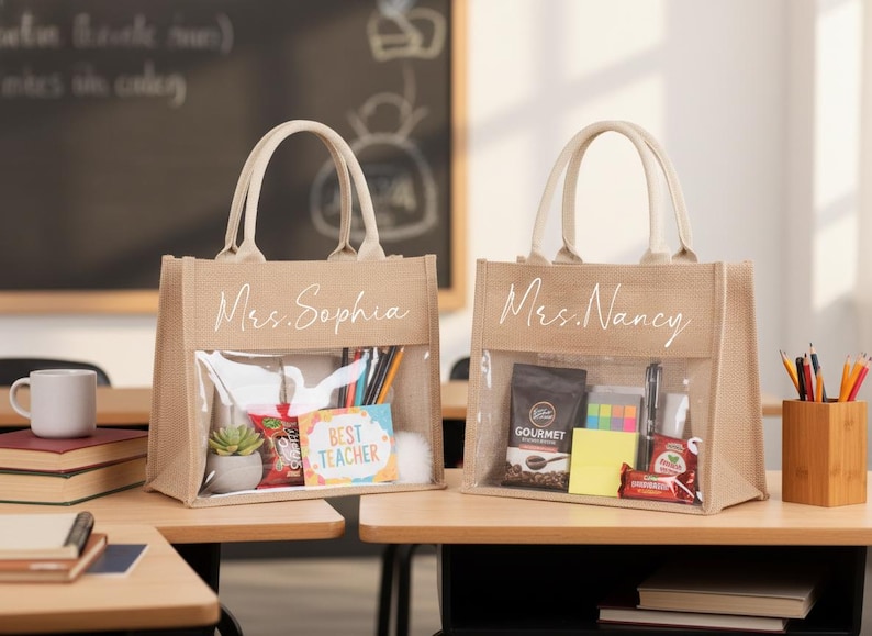May include: Two beige tote bags with clear front pockets, filled with gifts. One bag says "Mrs. Sophia" and the other says "Mrs. Nancy" in white script. The bags sit on a wooden desk, with a chalkboard in the background.
