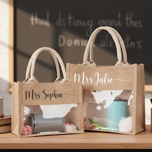 May include: Two burlap tote bags with clear front panels. The bags have white handles and the names "Mrs. Sophia" and "Mrs. Julie" printed on them. One bag contains a tumbler and flowers.