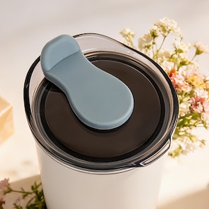 May include: A white travel mug with a black interior and a light blue lid. The lid has a flip-top closure. The mug is partially visible, with a bouquet of flowers in the background.