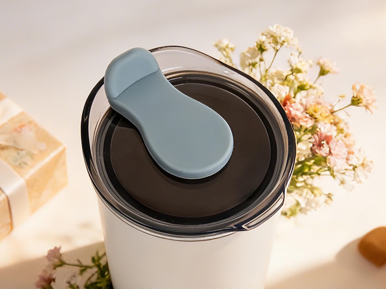 May include: A white travel mug with a black interior and a light blue lid. The lid has a flip-top closure. The mug is partially visible, with a bouquet of flowers in the background.