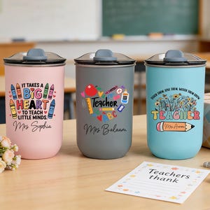 May include: Three insulated tumblers with lids, each featuring a teacher-themed design. One is pink with a crayon graphic and the text "It takes a big heart to teach little minds." The second is gray with a heart graphic and the word "Teacher." The third is blue with a floral graphic and the word "Teacher."