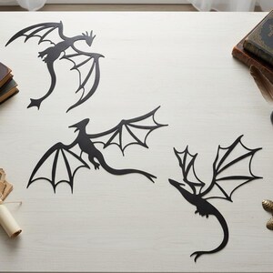 May include: Three black metal dragon wall art pieces with outstretched wings. The dragons have detailed wing designs and curved tails. The art pieces are displayed on a light-coloured surface, suggesting a fantasy or medieval theme.