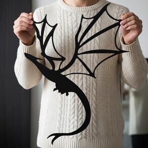 May include: A black metal dragon silhouette with outstretched wings. The dragon is held up by a person wearing a cream-coloured cable knit jumper. The dragon's wings are detailed with multiple points and lines.