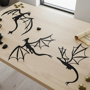 May include: Three black dragon silhouettes are displayed on a light wood surface. The dragons have detailed wings and varied poses. A rolled parchment with text is visible in the background. The dragons are a decorative item.