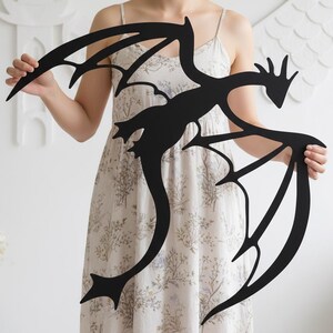 May include: A black metal dragon wall art piece with outstretched wings and a long, curved tail. The dragon silhouette is held up by a person. The dragon's wings have a detailed, stylized design.
