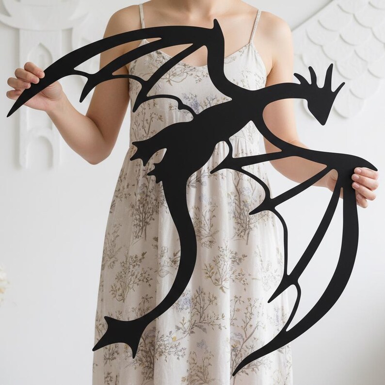 May include: A black metal dragon wall art piece with outstretched wings and a long, curved tail. The dragon silhouette is held up by a person. The dragon's wings have a detailed, stylized design.