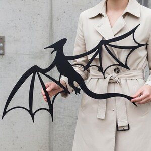 May include: A black metal dragon silhouette with outstretched wings. The dragon has a long tail and a detailed body shape. The wings have a stylized, pointed design. The dragon is held in front of a neutral background.