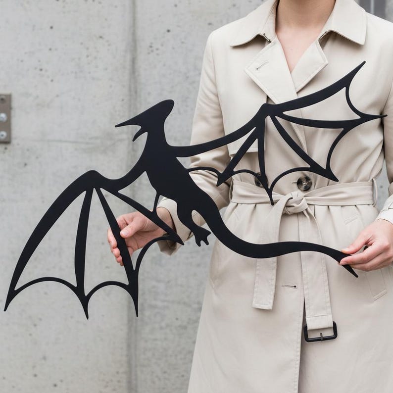 May include: A black metal dragon silhouette with outstretched wings. The dragon has a long tail and a detailed body shape. The wings have a stylized, pointed design. The dragon is held in front of a neutral background.
