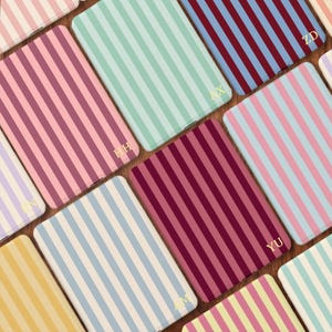 May include: A collection of rectangular cases featuring vertical stripes in various color combinations including pink, mint green, burgundy, blue, and yellow. Each case displays two gold-colored capital letters embossed in the bottom corner.