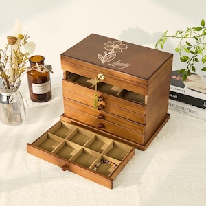 Może przedstawiać: Drewniane pudełko na biżuterię z wieloma szufladami i otwieraną pokrywą. Pudełko jest bejcowane na ciepły brąz i posiada kwiatowy wzór oraz napis "Luxury" na pokrywie. Szuflady wyłożone są beżową tkaniną, a ze zamka zwisa złoty frędzel.