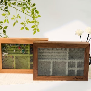 Op de afbeelding: Twee houten sieradendoosjes met glazen voorkant. Eén doos is lichtgekleurd en toont sieraden op een groene fluwelen binnenkant. De andere is donker gekleurd met een patroon glazen voorkant en de naam "Blakely" rechtsonder.