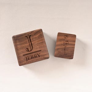 May include: Two wooden coasters. One square coaster features the letter "J" above the name "JERRY". The other rectangular coaster has a floral design and the name "Laurel". Both coasters are made of dark wood.