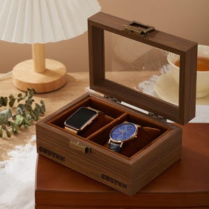 May include: A wooden watch box with a clear glass lid, open to show two compartments lined with brown velvet. One holds a gold smartwatch, the other a blue-faced watch with a black leather strap. The box is engraved with the word "CUSTOM".