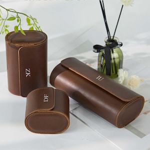 May include: Three brown leather watch cases of varying sizes. Each case has a flap closure and a personalized monogram in white. The cases are cylindrical and oval-shaped, with a smooth, polished finish. The cases are displayed on a white surface.