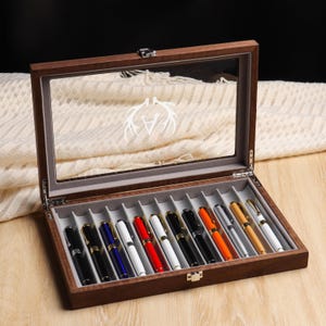 May include: A wooden pen display case with a glass top, showcasing a collection of colorful pens. The case is open, revealing pens in various colors, including black, blue, red, and gold. The interior is lined with gray fabric.