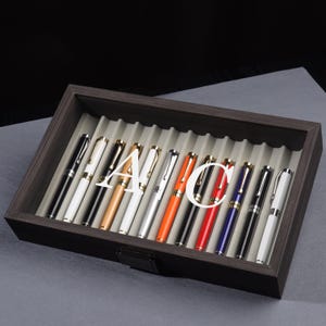Puede incluir: Un estuche de madera para bolígrafos con tapa de cristal, que muestra una colección de plumas estilográficas. Los bolígrafos son de varios colores, incluyendo negro, blanco, dorado, plateado, naranja, rojo y azul. El estuche está forrado con fieltro gris.