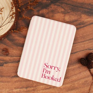Puede incluir: Una funda rectangular para lector electrónico con un patrón de rayas verticales en rosa claro y blanco. El texto Sorry, I'm Booked está impreso en una fuente negrita de color rosa oscuro en la esquina inferior derecha de la cubierta.