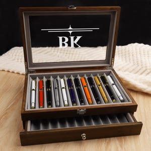May include: A wooden pen display case with a glass top, showcasing a collection of colorful pens. The case has a drawer below and the initials "BK" are displayed on the glass. The pens are in various colors, including white, red, black, and blue.
