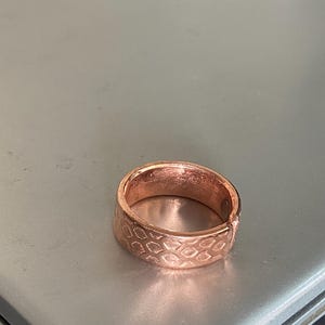May include: A copper-colored ring with a textured, geometric pattern. The ring has a band design with a series of connected shapes. The ring is made of a shiny metal and is displayed on a gray surface.