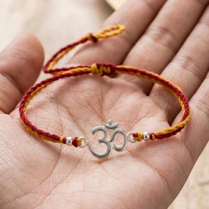 May include: A bracelet featuring a silver-toned Om symbol charm centered on a braided cord made of intertwined red and yellow threads. The design includes small silver-toned beads flanking the central charm and an adjustable sliding knot closure.