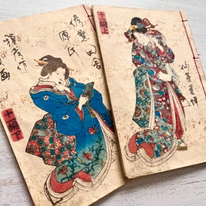 Op de afbeelding: Twee vintage Japanse houtsnede boeken met illustraties van figuren in traditionele gewaden met patronen. Het linkerboek toont een figuur in een blauw gewaad die een voorwerp vasthoudt, terwijl het rechterboek een staande figuur in een bloemrijk gewaad toont.