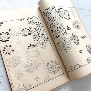 May include: An open book with cream-colored pages featuring black ink illustrations. The left page displays a child figure and various floral motifs. The right page contains multiple grid-like sections filled with intricate botanical patterns and Japanese text.