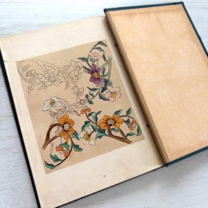 以下が含まれることがあります： ダークティール色の背表紙とクリーム色のページを持つ、開かれたハードカバーの本。左側のページには、オレンジ、紫、白、緑の色合いで描かれた様式化された花の植物画が詳細に記されており、一部は線画のままになっています。