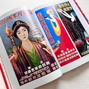 May include: An open book displaying three vintage-style posters with Japanese text. The left poster features a person holding a flaming torch, the center shows a figure in a blue and yellow robe, and the right depicts a person in a black dress.