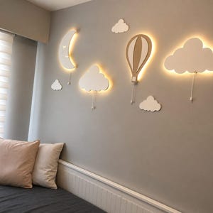 Könnte beinhalten: Eine skurrile Wanddekoration mit einem Halbmond, einem Heißluftballon und wolkenförmigen Lichtern. Die Lichter sind weiß mit einem warmen Schein, der eine sanfte Atmosphäre schafft. Die Dekoration ist an einer grauen Wand angebracht, mit Kissen und einem Bett im Vordergrund.