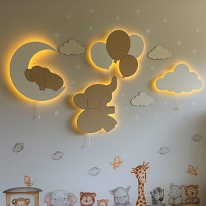 Op de afbeelding: Een grillige wanddecoratie voor de kinderkamer met een slapende olifant op een maan, een olifant die ballonnen vasthoudt en wolkenaccenten. De houten uitsparingen zijn verlicht met warme LED-lampen, waardoor een zachte gloed ontstaat. Extra dierenfiguren zijn hieronder geplaatst.