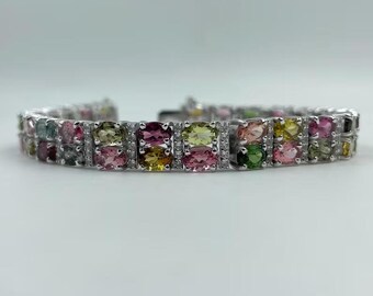 May include: A silver bracelet featuring a row of colorful gemstones. The gemstones are a mix of pink, green, yellow, and other colors. The bracelet is set against a white background.