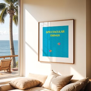 May include: A framed art print with the words "SPECTACULAR THINGS" in yellow text on a blue background. Two fish swim in the water. The print is in a wooden frame and hangs on a white wall near a window overlooking the ocean.