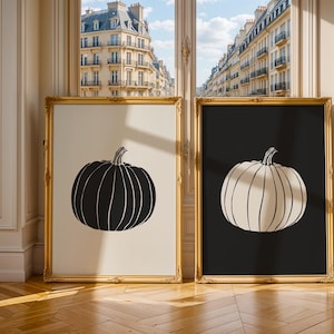 Puede incluir: Dos impresiones artísticas enmarcadas con ilustraciones de calabazas. Una presenta una calabaza negra sobre fondo crema, la otra una calabaza crema sobre fondo negro. Ambas en marcos dorados.
