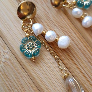 Könnte beinhalten: Goldfarbene Ohrringe mit floralem Design und hängenden Elementen. Jeder Ohrring hat einen goldenen Stecker, eine türkisfarbene Blume, weiße Perlen, eine goldene Kette und eine transparente, ovale Perle. Die Ohrringe sind vor einem Holzgrund platziert.