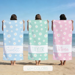 May include: Three personalized beach towels in blue, mint green, and pink, each adorned with white daisy patterns. The towels feature a white band with names: Scarlett, Taylor, and Victoria. The image also includes the text "40+ COLORS".