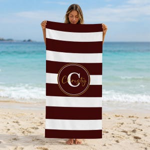 May include: A rectangular beach towel featuring horizontal stripes in deep maroon and white. Centered on the towel is a circular emblem with a gold outline, containing a large white letter C and the name Christy written in gold script.