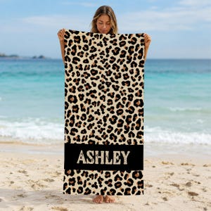 May include: A rectangular beach towel featuring a repeating leopard print pattern in shades of tan, brown, and black. A solid black horizontal band is positioned near the bottom, displaying the name ASHLEY in a distressed white serif font.