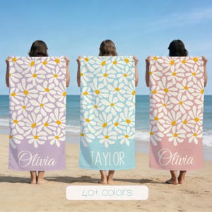 May include: Three beach towels with daisy patterns in lavender, light blue, and pink. Each towel has a personalized name at the bottom: Olivia and Taylor. The towels are held up on a beach with the ocean in the background. The text "40+ colors" is at the bottom.