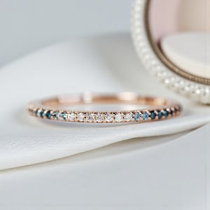 May include: A rose gold ring featuring a band of small, sparkling white and blue gemstones. The ring is displayed on a white surface, with a blurred decorative element in the background.