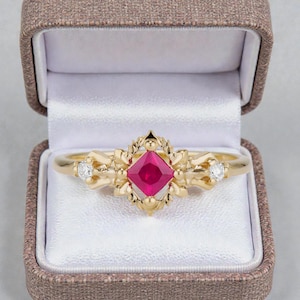 May include: A gold ring with a square, red gemstone at the center, flanked by two smaller white gemstones. The ring is displayed in an open, brown jewelry box with a white satin interior. The ring's design features ornate floral details.