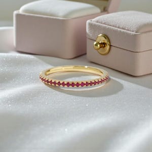 May include: A gold ring featuring a continuous line of small, red gemstones. The ring is set against a white, textured backdrop, with two pink jewellery boxes in the background.