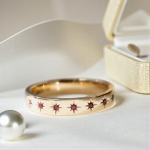 Könnte beinhalten: Ein goldener Bandring mit Sternmustern, die jeweils einen kleinen roten Edelstein enthalten. Der Ring wird auf einer weißen Oberfläche präsentiert, mit einer Perle und einer Schmuckschatulle im Hintergrund. Das Design ist elegant.