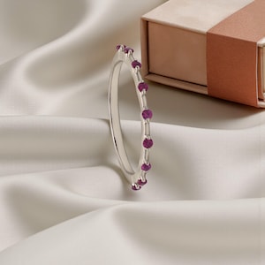 Può includere: Un anello d'argento con una fila di piccole pietre preziose rotonde color magenta. L'anello è posto su uno sfondo di tessuto bianco e una scatola regalo parzialmente visibile con un nastro marrone.