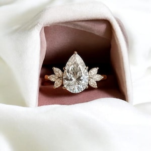 Puede incluir: Un anillo de oro con un gran diamante en forma de pera como pieza central. El anillo está flanqueado por diamantes más pequeños en forma de pétalo a cada lado, sobre un fondo de terciopelo rosa y tela blanca.