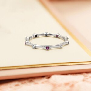 Könnte beinhalten: Ein silberner Ring mit einem Bambus-inspirierten Design. Der Ring hat abwechselnd Edelsteine, darunter rote, schwarze und klare Steine. Der Ring wird auf einer hellen Oberfläche präsentiert, die seine komplizierten Details und seinen eleganten Stil hervorhebt.