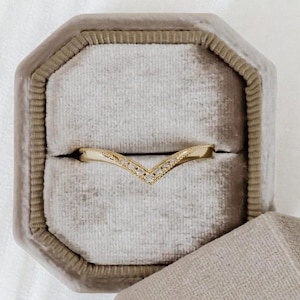 Pode incluir: Um anel dourado em forma de V com pequenas pedras preciosas transparentes colocadas ao longo da curva superior. O anel é exibido dentro de uma caixa de anel de veludo cinza com formato octogonal.