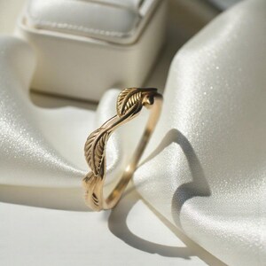 May include: A gold-colored ring with a leaf design. The ring features a delicate, intertwined leaf pattern. The ring is displayed on a white satin fabric, with a white jewelry box in the background. The ring is a piece of jewelry.