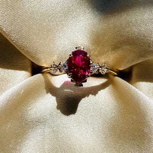 May include: A gold ring with a large, oval-cut, deep red gemstone as the centerpiece. The ring is adorned with smaller, clear gemstones on either side of the main stone, set against a cream-colored fabric.